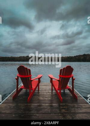 Deux chaises rouges adirondack à l'extrémité du quai sur le lac le jour de tempête. Banque D'Images