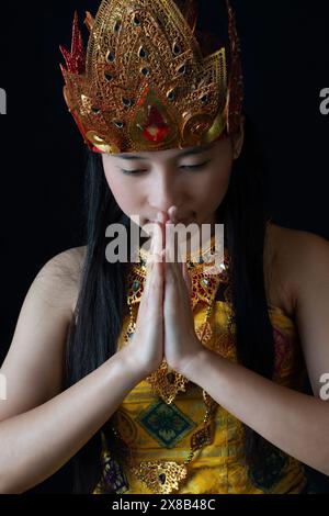 Une femme indonésienne vêtue d'une tenue dorée traditionnelle avec un couvre-chef orné, capturée dans une pose de prière Banque D'Images