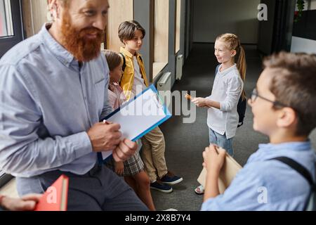 Un enseignant masculin tenant un presse-papiers se tient à côté d'un groupe d'enfants dans un cadre de classe dynamique. Banque D'Images