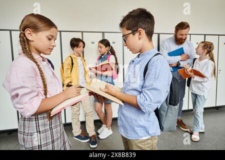 Un groupe diversifié de jeunes enfants, dirigé par un enseignant masculin, se tiennent autour les uns des autres dans une salle de classe lumineuse et animée. Banque D'Images