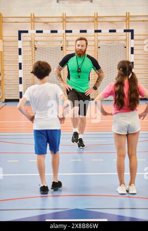 Un groupe diversifié d'enfants, dirigé par un enseignant masculin, s'exerçant ensemble sur un sol de salle de sport très éclairé. Banque D'Images