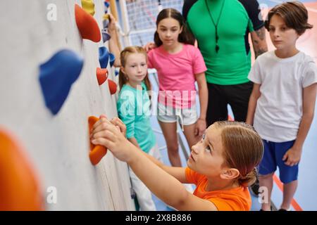 Un groupe diversifié de jeunes enfants se tiennent ensemble, écoutant attentivement leur professeur masculin dans une salle de sport lumineuse et animée. Banque D'Images