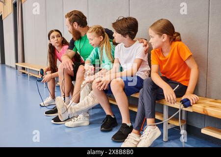 Un groupe diversifié de jeunes enfants, assis sur un banc, écoutant attentivement pendant que leur enseignant masculin transmet des connaissances dans un cadre de classe dynamique. Banque D'Images