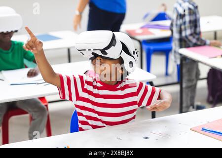 Dans la salle de classe de l'école, garçon biracial portant un casque de réalité virtuelle pointant vers le haut Banque D'Images