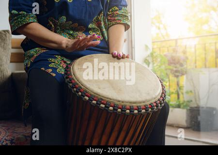 Mains féminines d'une femme méconnaissable jouant du tambour Djembe à la maison. Le tambour est marron et a un motif rouge et noir. La femme est dans un a détendu Banque D'Images