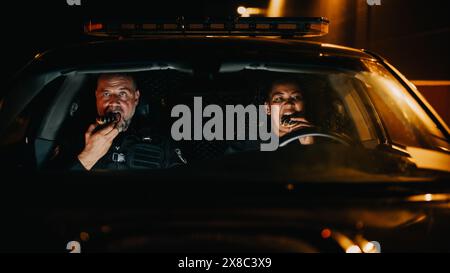 Deux policiers assis dans la voiture de patrouille de la circulation, mangeant des Donuts. Les partenaires de la police profitent de leur pause calme dans les rues paisibles. Service de nuit des forces de l'ordre. Point de vue de l'extérieur de la voiture Banque D'Images