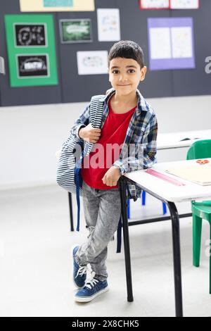 À l'école, un jeune garçon biracial avec un sac à dos se tient près d'un bureau dans la salle de classe Banque D'Images