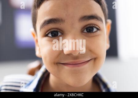 À l'école, un jeune garçon biracial aux cheveux bruns courts et aux yeux bruns sourit dans la salle de classe Banque D'Images