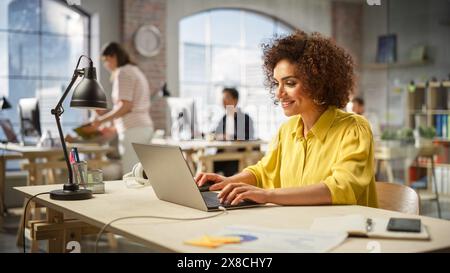 Portrait de jeune femme productive biraciale travaillant sur ordinateur dans le bureau occupé moderne. Chef d'équipe féminin souriant après avoir accompli une tâche importante. Les gens travaillant en arrière-plan. Banque D'Images