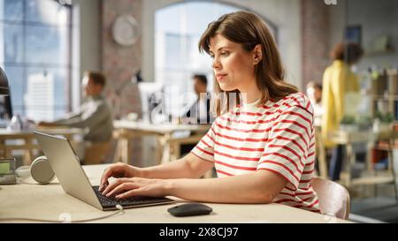 Portrait de femme caucasienne tapant sur un ordinateur portable au travail dans un bureau moderne spacieux. Responsable des opérations féminin vérifiant l'état du processus. Employés travaillant en arrière-plan. Banque D'Images