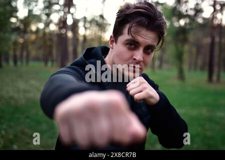 jeune homme sportif dans une veste noire sportive dans une pose de combat levant le poing pour un coup de poing sur un fond d'arbres forestiers Banque D'Images