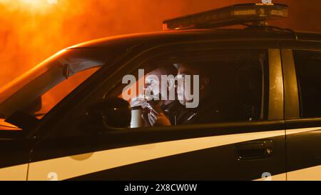 Portrait de deux officiers de police assis dans la voiture mangeant des Donuts, surveillant les rues pendant Misty Night. Officiers professionnels au repos de leur quart de travail. Partenaires policiers prenant une pause Banque D'Images