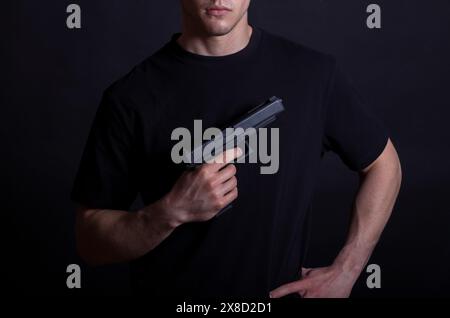 Jeune homme dans un t-shirt noir tenant un pistolet sur un fond sombre Banque D'Images