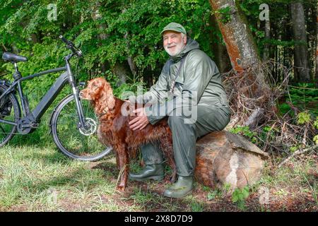 Un homme plus âgé prend une pause à la lisière de la forêt pendant sa balade à vélo. Son chien bien-aimé est assis entre ses jambes. Banque D'Images