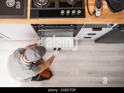 Un homme portant des vêtements de travail se concentre sur la fixation d'un poêle dans une cuisine moderne. Des outils et des pièces sont dispersés autour de lui pendant qu'il travaille avec diligence pour réparer le Banque D'Images