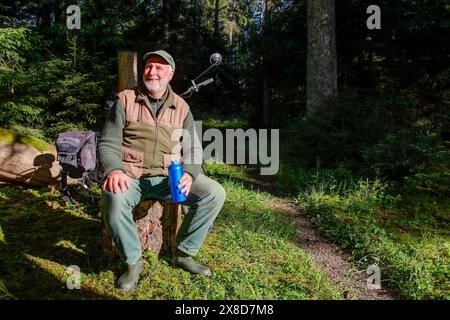 Un cycliste âgé repose sur une souche d'arbre dans la forêt, entouré par le soleil du matin et le silence, un moment de sérénité et de connexion à la nature. Banque D'Images