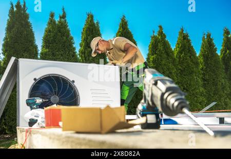 Un technicien CVC portant un chapeau et un pantalon vert travaille avec diligence sur un climatiseur ou une pompe à chaleur à l'extérieur. Banque D'Images
