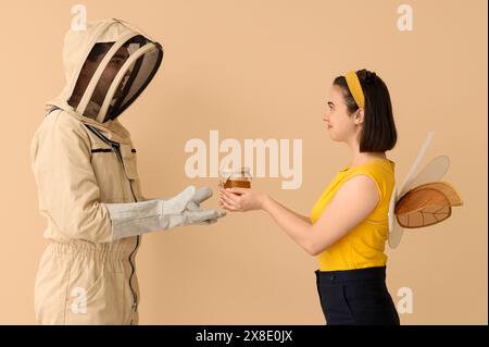 Apiculteur mâle avec pot de miel et femme en costume d'abeille sur fond beige Banque D'Images