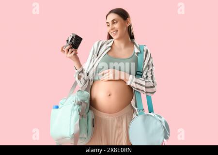 Jeune femme enceinte avec sacs d'hôpital de maternité et appareil photo sur fond rose Banque D'Images