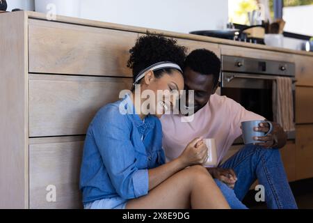 Un couple diversifié à la maison, assis dans la cuisine, prenant un café. Porter des vêtements décontractés, partager des moments joyeux, profiter de la compagnie de l'autre, inchangés Banque D'Images