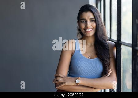 Dans un bureau moderne, une jeune femme sud-indienne sourit par une fenêtre, bras croisés, espace copie. Elle a de longs cheveux foncés, la peau brun clair, et porte Banque D'Images