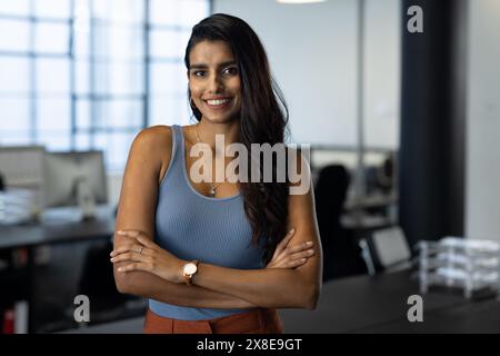 Au bureau moderne, la femme indienne se tient souriante, les bras croisés, en haut bleu sans manches. Elle a de longs cheveux foncés, des yeux bruns et un pantalon orange, avec calme Banque D'Images