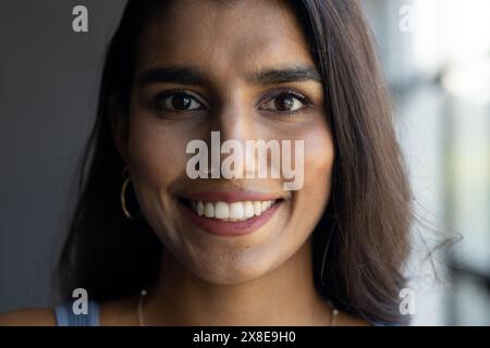 Dans un bureau moderne, une jeune femme indienne aux longs cheveux foncés et aux yeux bruns sourit chaleureusement. Porter des boucles d'oreilles, top bleu clair, maquillage naturel, et Banque D'Images