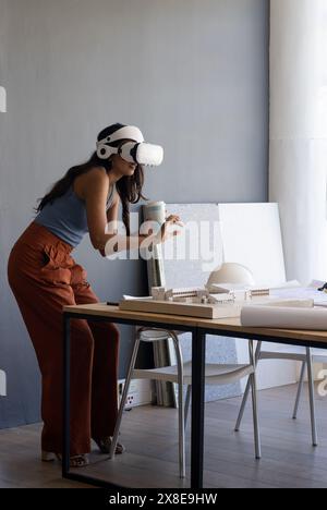 femme indienne, d'âge moyen, portant un casque de réalité virtuelle, debout dans un bureau, espace de copie. Tenant la tasse et regardant le modèle architectural sur la table, Banque D'Images
