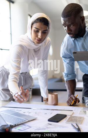 Au bureau moderne, équipe diversifiée travaillant sur des plans architecturaux. Jeune homme afro-américain avec les cheveux courts et la barbe, et jeune équipe féminine indienne lea Banque D'Images