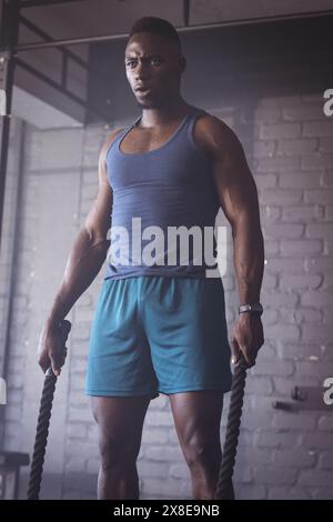 Au gymnase, homme afro-américain fort et en forme tenant des cordes de combat. Debout devant le fond de brique avec l'équipement de gymnastique autour, prêt pour w intense Banque D'Images