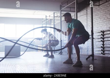 Deux hommes biraciaux utilisant des cordes de combat au gymnase, entourés par des hommes divers qui travaillent, copiez l'espace. Grandes fenêtres et divers appareils de fitness créant en Banque D'Images