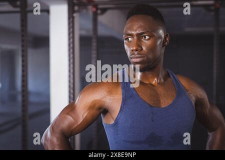 Homme afro-américain fort et en forme au gymnase dans le débardeur bleu, regardant déterminé. Debout dans une salle de gym moderne avec divers équipements d'entraînement en arrière-plan, Banque D'Images
