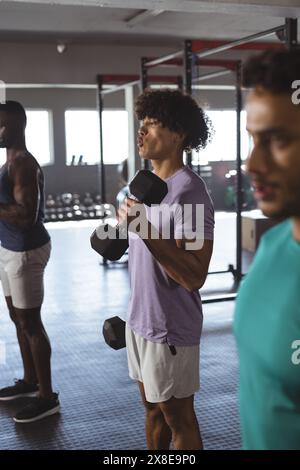 Au gymnase, trois hommes s'adaptent à de jeunes hommes divers qui s'entraînent avec des haltères. Ils sont concentrés sur leur entraînement dans une salle de gym spacieuse et bien éclairée avec divers équipements dans bac Banque D'Images