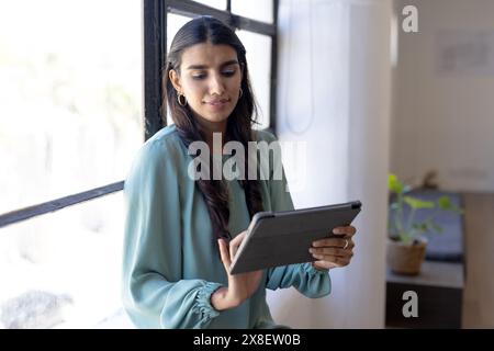 Dans le bureau moderne, jeune femme indienne avec de longs cheveux foncés et boucles d'oreilles par fenêtre. Elle porte un chemisier bleu clair, regardant l'écran soigneusement Banque D'Images