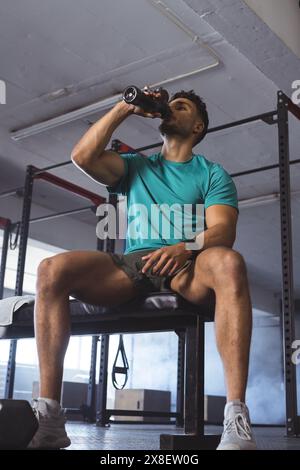 Au gymnase, jeune homme biracial amateur de fitness boire de l'eau après l'entraînement. Équipement de gymnastique et poids visibles en arrière-plan, créant une forme physique ciblée Banque D'Images