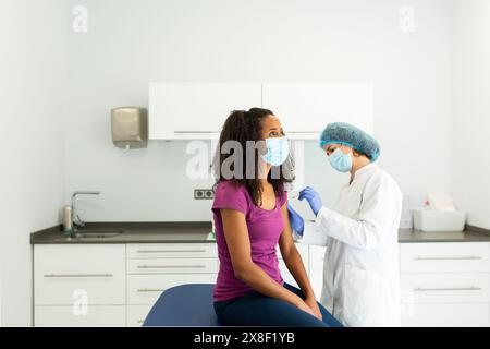 Médecin se préparant à administrer une injection de vaccin à un patient assis sur une table d'examen. Les deux individus portent des masques et sont engagés dans un convers Banque D'Images