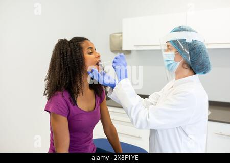 Travailleur de la santé administrant un test COVID-19 en prélevant un échantillon de prélèvement dans la bouche d'un patient. Patient assis alors qu'un travailleur de santé porte du pr Banque D'Images