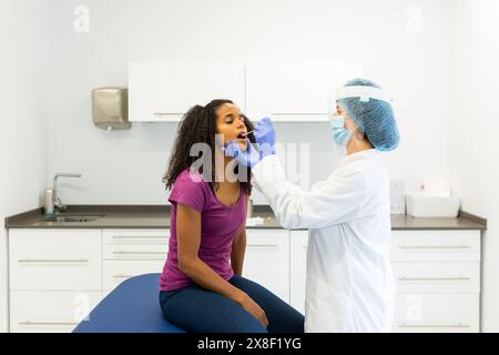 Travailleur de la santé administrant un test COVID-19 en prélevant un échantillon de prélèvement dans la bouche d'un patient. Patient assis alors qu'un travailleur de santé porte du pr Banque D'Images