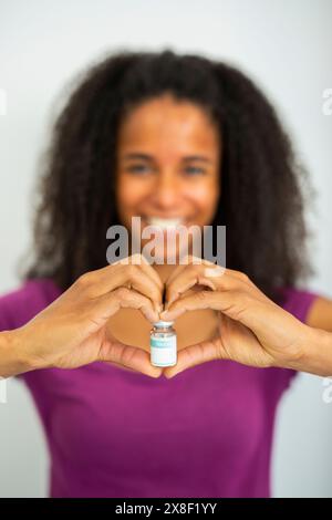 Portrait d'une femme souriante tenant un flacon de vaccin entre ses mains, formant une forme de coeur. Banque D'Images