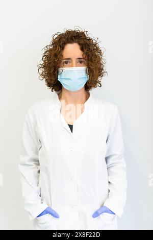 Portrait d'un travailleur de la santé portant un masque de protection bleu. Docteur portant un manteau blanc debout contre un fond blanc et regardant directement Banque D'Images