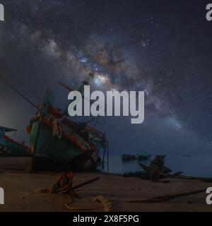 Vue astronomique du ciel avec voie lactée au premier plan, mer, plage et vieux bateaux de pêche garés sur la plage de Koh Chang, Thaïlande. Banque D'Images