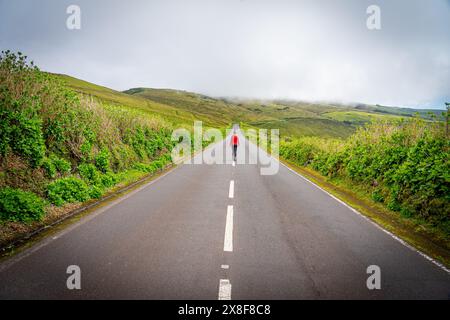 Femme méconnaissable marchant au milieu de la route entre les lignes pointillées habillées de rouge parmi la végétation verte. São Jorge Island-Açores Banque D'Images