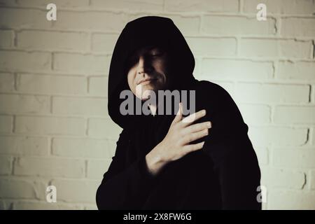 Un homme portant un sweat à capuche noir danse énergiquement avec des mouvements expressifs de la main devant un mur de briques blanches. Banque D'Images