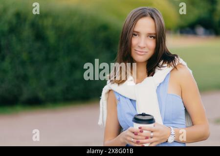 Femme confiante avec une tasse de café pose sur le fond luxuriant d'un parc, son regard serein et engageant. Banque D'Images