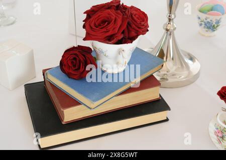 Une pile de trois livres avec une tasse à thé remplie de roses rouges sur le dessus, placée sur une nappe blanche. Banque D'Images