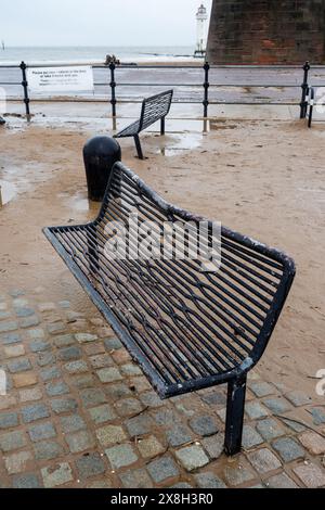 Bancs en métal noir vides sur une promenade de plage pluvieuse avec du sable humide et un ciel nuageux en arrière-plan. Banque D'Images