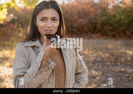 Femme millénaire hispanique dictant un message audio sur smartphone marchant dans le parc d'automne Banque D'Images