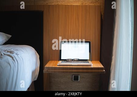 Un ordinateur portable avec des lunettes sur le clavier placé sur un petit bureau en bois à côté d'un lit dans une pièce confortable. Banque D'Images