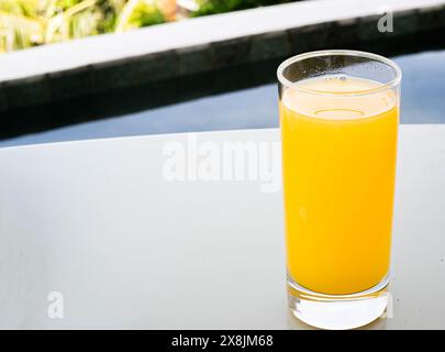 Un verre de jus d'orange placé sur une surface blanche près d'une piscine avec de la verdure en arrière-plan. Banque D'Images