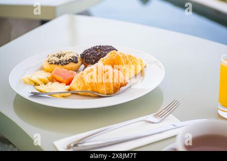 Une assiette de petit déjeuner avec croissants, beignets, tranches d'ananas et un morceau de saumon, accompagné d'un verre de jus d'orange et d'une tasse de café sur a ta Banque D'Images